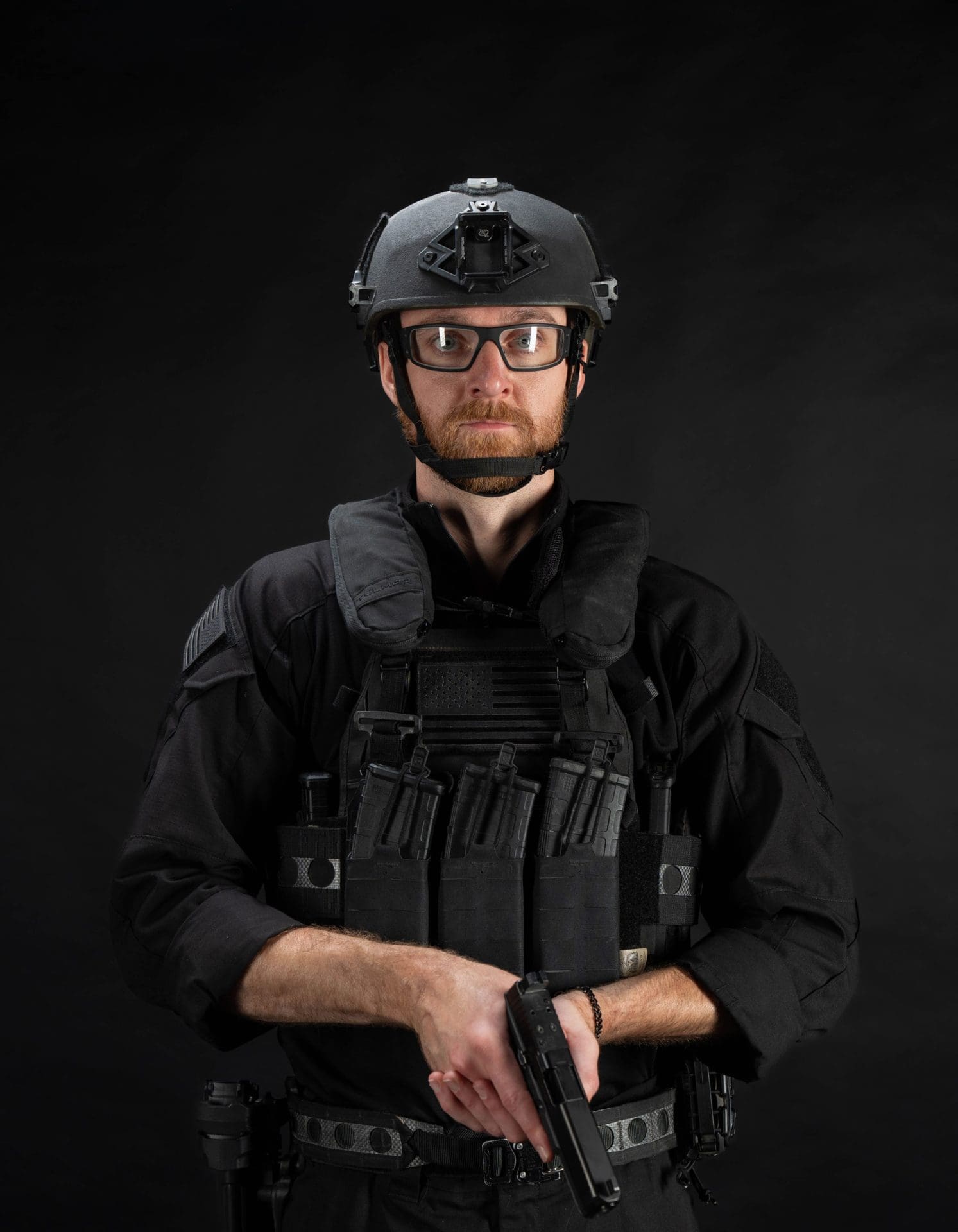Person in tactical gear holding a firearm against a black background.