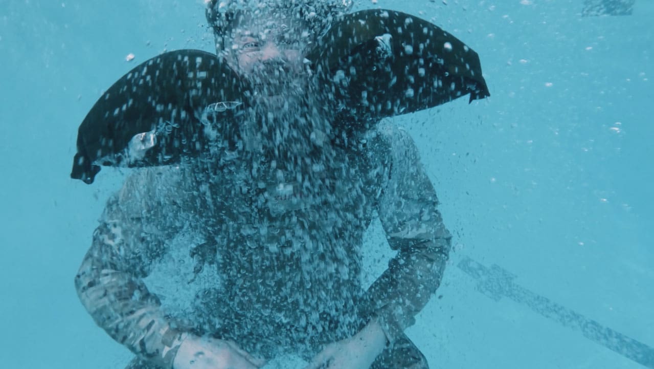 Person submerged in water, bubbles surrounding the body, wearing a dark outfit and the inflated Galeo around his neck.